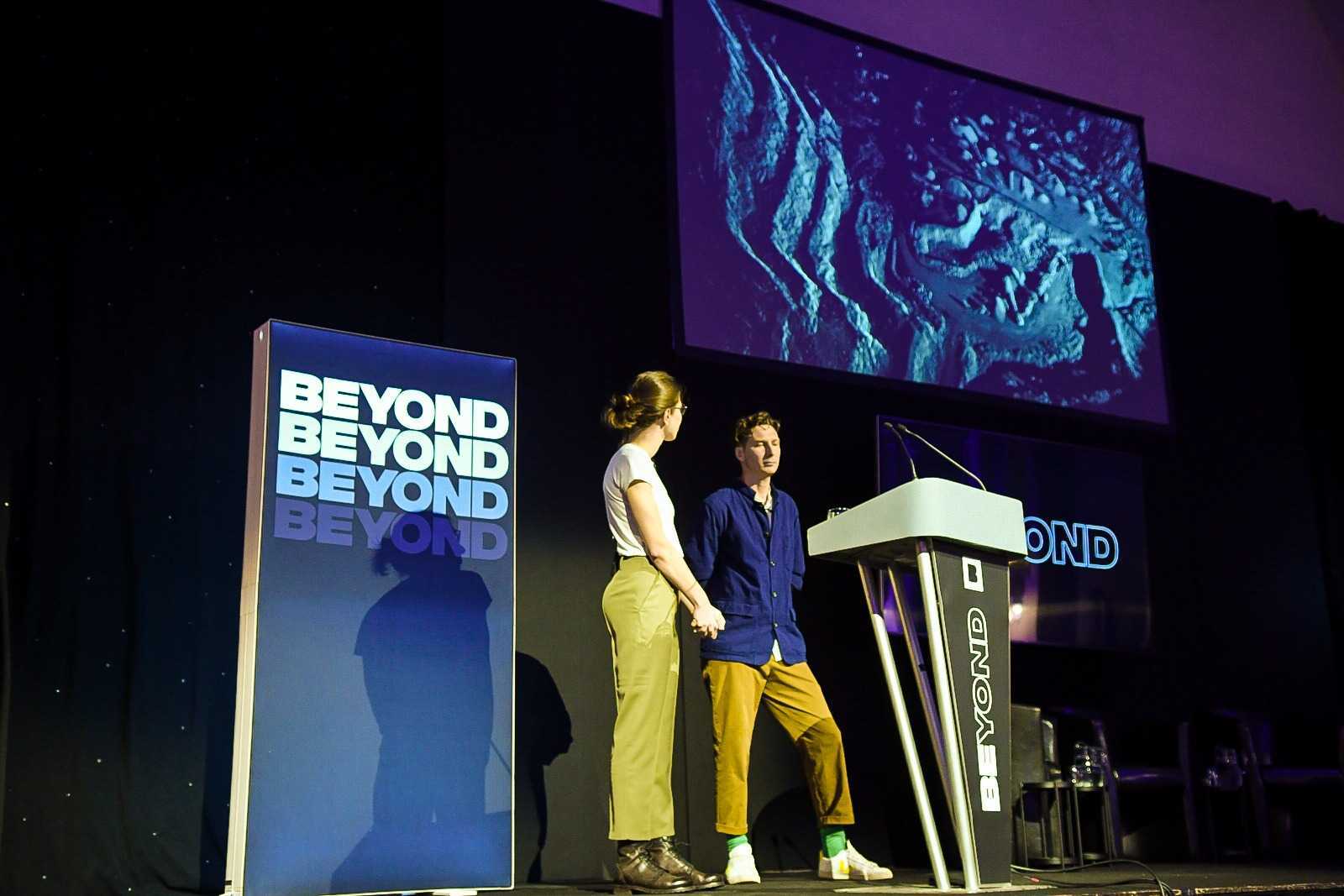 two speakers on stage at Beyond conference. A woman wearing green trousers and a white top is looking at a man wearing yellow trousers and a navy shirt. They are stood behid a speaker podium with a large screen above them