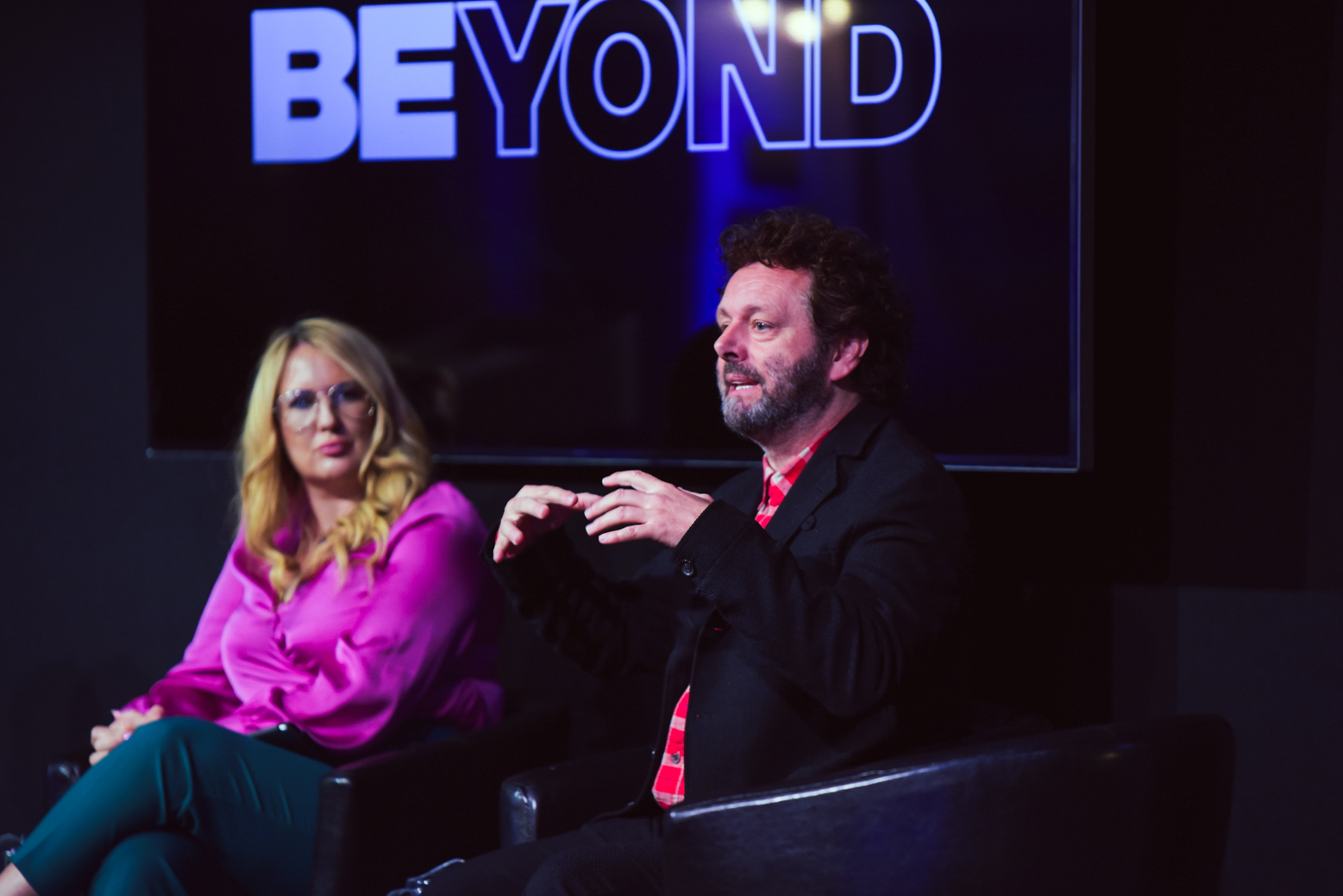Sophie Howe and Michael Sheen on stage at Beyond conference. They are both sitting down. Michael is mid-speech, Sophie is looking at him